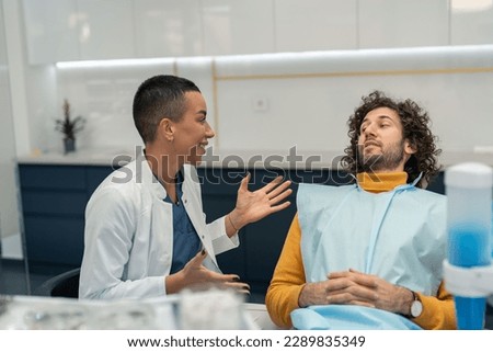 Young woman dentist sitting in her dental care office next to male patient on dentist's chair, recommending and explaining dental procedures with smile, motivating patient before treatment.