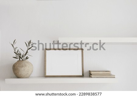 Elegant interior still life. Two floating shelves. Blank wooden picture frame mockup template. Textured vase with olive tree branches and old books. Modern Mediterranean home. White wall background.