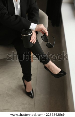 a girl sitting on an armchair in a black suit with a white blouse on her feet high-heeled shoes and leggings in her hands a thermos mug and glasses Royalty-Free Stock Photo #2289842569