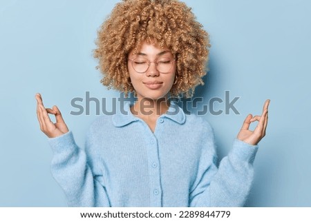 Stress relief concept. Curly haired calm woman meditates with closed eyes practices yoga searches for inner balance wears spectacles soft jumper isolated over blue background. Calmness and mindfulness
