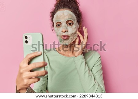 Skin care procedures. Lovely darkhaired woman applies facial bubble mask for pampering keeps lips rounded poses for making selfie or records video for her beauty blog poses against pink background Royalty-Free Stock Photo #2289844813