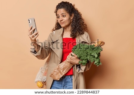 Displeased curly haired young Brazilian woman takes selfie via smartphone poses with grocery products has shopping day wears jacket and jeans isolated over brown background bought organic food