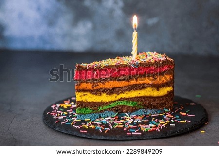 Rainbow birthday cake decorated with a festive candle and confetti. Confectionery. Multicolored dessert in the color of the rainbow on a black background.