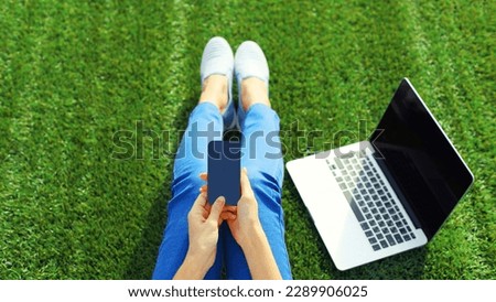 Modern woman holding smartphone with laptop in summer park sitting on green grass