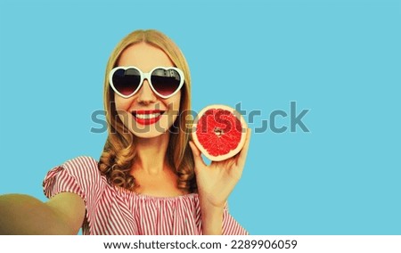 Summer portrait of happy smiling young woman posing stretching her hand for taking selfie with smartphone with juicy slice of grapefruit fruit wearing heart shaped sunglasses on blue background