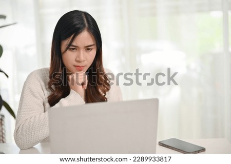 Thoughtful and focused young Asian woman looking at her laptop screen, working on her tasks on laptop at home. work from home concept