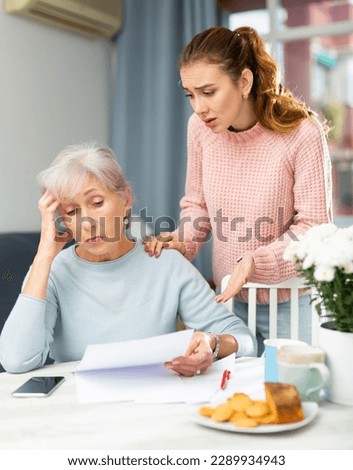 Portrait of elderly woman and her adult daughter looking at papers at home, worrying about debts on bills.