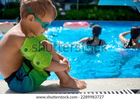 Frustrated boy in bathing shorts and glasses is sitting on the curb by the pool, hugging his knees. Sad child in swimming gear watches adults swimming in a deep pool