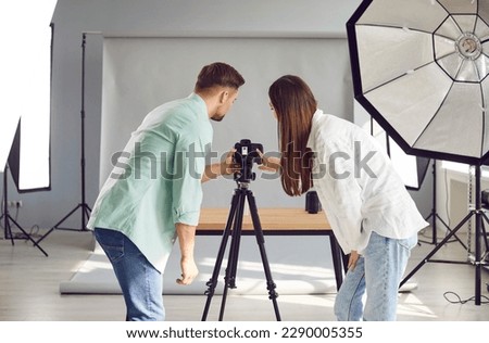 Team of two professional photographers working in their photo studio. Back view of young man and woman standing by tripod with camera and looking at object on table. Professional photography concept