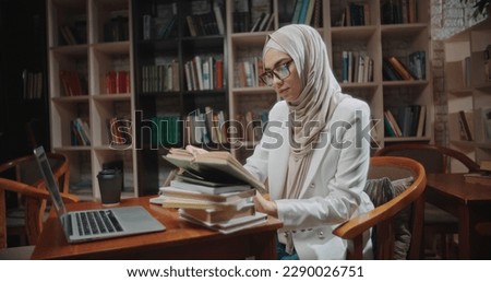 Muslim student is doing research at desk full of books in library. Girl wearing hijab is preparing for exam or making a project - student lifestyle, modern Islam close up 