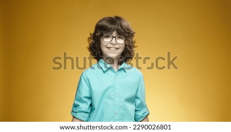 Little caucasian boy in blue shirt and glasses looking at camera, positively smiling, isolated on yellow background - emotions concept close up 