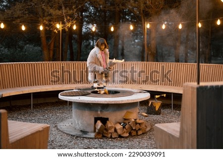 Young woman prepares food on beautiful outdoor lounge area with fire and round bench in pine forest at dusk. Luxury lifestyle at countryside concept