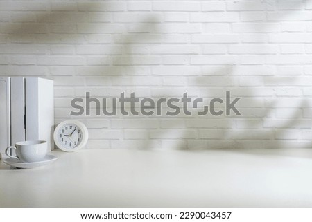 Minimal workplace with books, clock and cup of coffee on white table against brick wall.
