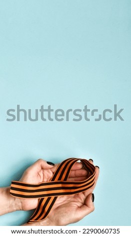 Hands holding george ribbon on blue background. Orange and black striped ribbon symbol on May 9. Victory day. Postcard for holiday. Vertical banner. Flat lay, copy space Royalty-Free Stock Photo #2290060735