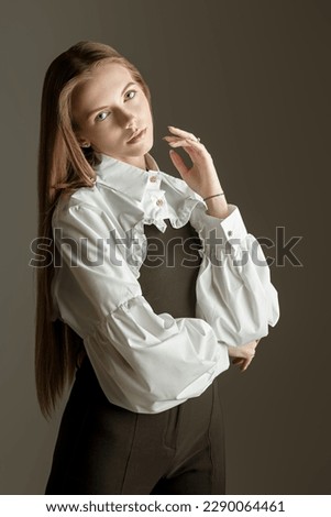 Beautiful girl model with long straight hair posing in a stylish white blouse, a black top and trousers on a gray studio background. Classic clothing style. Style and Fashion. 
