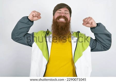Strong powerful red haired man wearing printed shirt over white studio background toothy smile, raises arms and shows biceps. Look at my muscles!