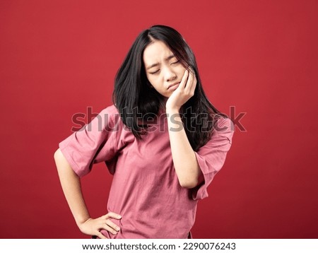 A portrait of a young Indonesian (Asian) woman wearing a pink shirt, looking like she has a toothache and holding her cheek. Isolated with a red background