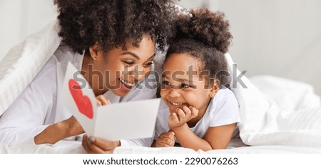 Cute little girl daughter congratulating his mom happy african american woman with Mother's day, giving her handmade greeting postcard with red heart at home. Family holidays concept
