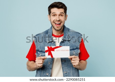 Young happy surprised man he wear denim vest red t-shirt casual clothes hold gift certificate coupon voucher card for store isolated on plain pastel light blue cyan background studio Lifestyle concept