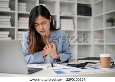 Unwell millennial Asian businesswoman feeling chest pain, suffering from heart attack during work at her desk in the office. Royalty-Free Stock Photo #2290100529