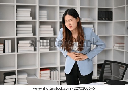 Sick and unwell millennial Asian businesswoman suffering from stomachache or abdominal pain in her office. Royalty-Free Stock Photo #2290100645
