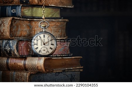 Old clock hanging on a chain on the background of old books.  Сlock as a symbol of time a books are a symbol of knowledge. Concept on the theme of history, nostalgia, culture. 