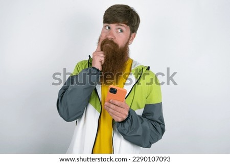 Image of a thinking dreaming red haired man wearing printed shirt over white studio background using mobile phone and holding hand on face. Taking decisions and social media concept.
