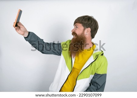 Portrait of a red haired man wearing printed shirt over white studio background  taking a selfie to send it to friends and followers or post it on his social media.