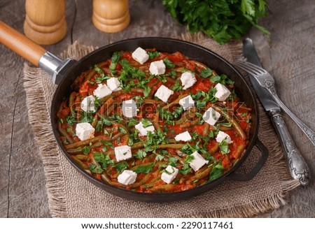Greek Green beans stew Fasolakia. Tender green beans braised in olive oil and tomato sauce served in cast iron skillet with feta cubes and fresh herbs. Selective focus, horizontal, wooden background.