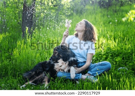 Young happy woman sitting on the grass with her dog blowing on dandelions on a summer day Royalty-Free Stock Photo #2290117929