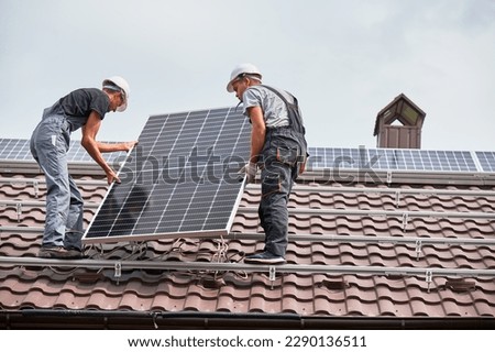 Men technicians lifting photovoltaic solar modul on roof of house. Electricians in helmets mounting solar panel system outdoors. Concept of alternative and renewable energy. Royalty-Free Stock Photo #2290136511