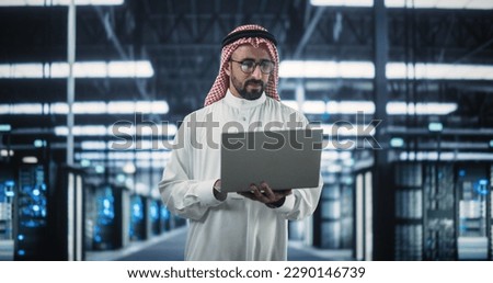 Portrait of a Middle Eastern Data Center Administrator Wearing Traditional White Clothes, Using Laptop Computer in a Modern Technological Server Farm Facility. Arab Cybersecurity Expert at Work Royalty-Free Stock Photo #2290146739