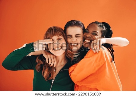 Three multicultural female friends smiling and embracing each other in a studio. Group of vibrant young women enjoying themselves while standing against an orange background. Royalty-Free Stock Photo #2290151039