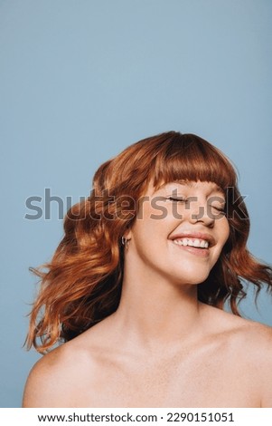 Woman with ginger hair and flawless skin smiling happily with her eyes closed. Confident young woman feeling comfortable in her own skin. Young woman standing topless against a blue studio background.