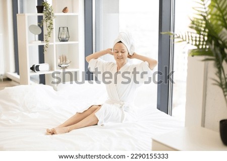 Relaxed young woman in robe and towel sitting barefoot with hands behind her head and closed eyes in bedroom. Serene caucasian lady enjoying leisure time near picture window on Sunday morning.