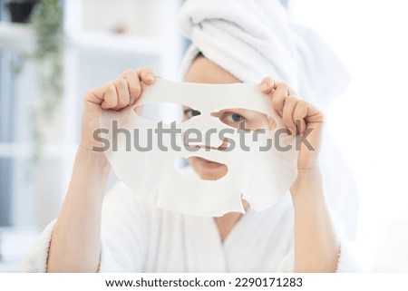Charming caucasian female in white robe and bath towel looking through holes of cosmetic sheet mask cut out for eyes on background of home interior. Pretty woman having fun with face care product. Royalty-Free Stock Photo #2290171283