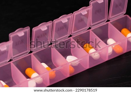 Close-up of a filled pink pillbox, an organizer for pills, vitamins and food additives on a black table, macro photography. Concept of maintaining health