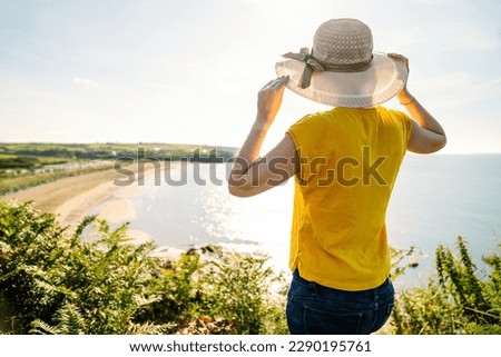 Vibrant Tourist: Back View of Colorful-Shirted Girl Touching Hat, Admiring Coastal Landscape from Cliffs in Noon Light