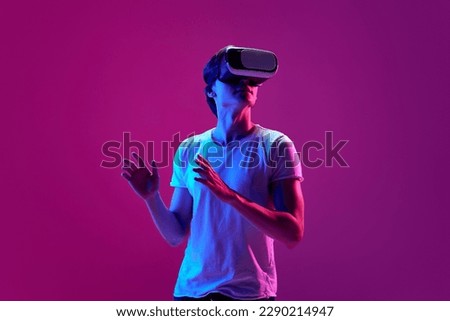 Escape from reality. One young guy wearing VR headset glasses over pink neon background. Youth and virtual lifestyle of future. Concept of games, modern, digitalization and technology Royalty-Free Stock Photo #2290214947