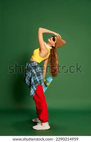 Photo of beautiful skateboarding girl wearing trend clothes ans sunglasses, holding skateboard in hands and posing over green background. Concept of youth, fashion, sport, activity and leisure time
