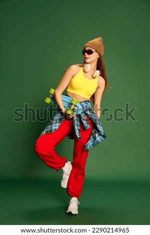 Full-length photo of skateboarding girl wearing stylish clothes and holding skateboard in hands over green background. Concept of youth, fashion, sport, hooby, fun, activity and leisure time