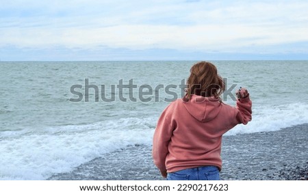 A girl in a pink hoodie throws pebbles into the sea, windy weather, pastel colors, waves, fingers, relaxation, melancholy, a leisurely walk, coast, cloudy, horizon line, redhead, seascape, emotions