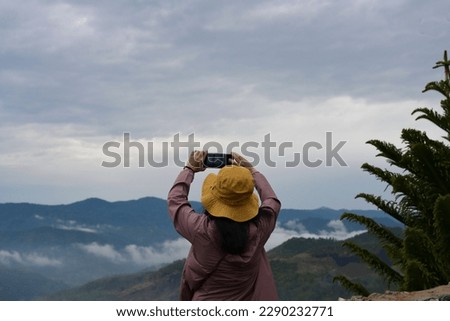 Woman wearing purple-brown robe wearing yellow wide-brimmed hat holds camera to take pictures in morning misty and dew. Tourists use small digital camera take pictures of fog-shrouded mountain views. 