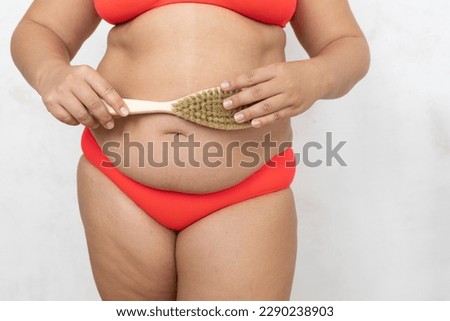 Cropped overweight, fat big female in red bikini scrubbing belly using brush with natural bristles, dry massage for healthy body and beauty. Big size dangling down belly problem, increase skin turgor