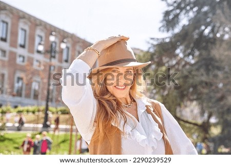  A carefree blonde woman with long hair and a stylish hat gazes directly into the camera, enjoying the peaceful surroundings of a park.