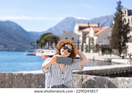Young smiling woman in a hat taking a selfie using a smartphone on the shore of a mediterranean sea village
