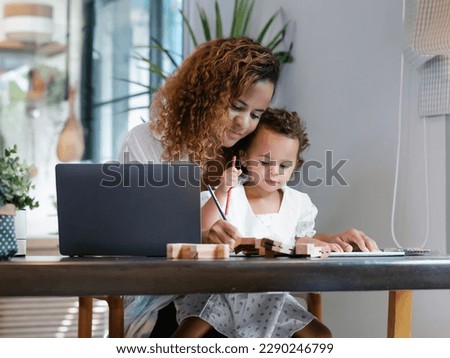 Smiling African American mom working with laptop computer while sitting wooden table in kitchen and playing with her baby girl. Young Black mother do freelance the desk with toddler child at home.