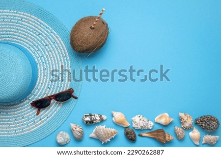 Summer and vacation flat lay with a beauty blue striped woman straw hat, sun glasses, a Coconut with straw and various seashells at the lower edge of the picture on blue background.