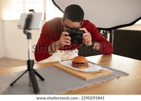 Food photographer taking photo of delicious juicy meaty hamburger in professional photo studio, brunette man wearing eyeglasses and casual outfit using digital camera and cell phone