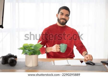 Portrait of cheerful happy handsome hispanic young man wearing stylish casual outfit photographer posing at workplace, guy sitting at desk with computer, tablet and notepad, drinking coffee